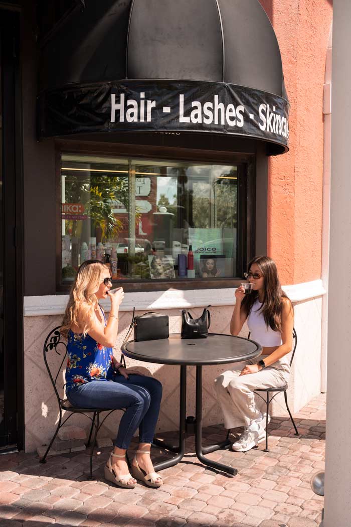 Two women sitting outside at a table drinking wine under the Beauty on Broadway "Hair - Lashes - Skincare" sign